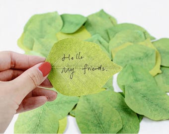 May include: A light green leaf-shaped sticky note with the text "Hello, my friends" in black script is held by a hand with red nail polish. The note is surrounded by other green leaf-shaped notes.