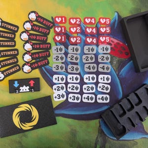 Peut inclure: Ensemble de jetons et de conteneurs de jeu. Comprend des jetons noirs avec le mot "STUNNED", "+1 BUFF" et d'autres symboles de jeu. Il y a aussi des jetons rouges et blancs avec des chiffres et des symboles de cœur, et des conteneurs noirs.