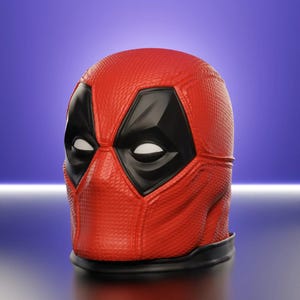 May include: A red Deadpool mask with black eye and mouth details. The mask has a textured surface and a black base. The eyes are white. The mask is a replica of the character from the Marvel comics.