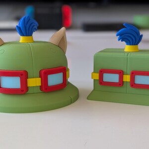 Puede incluir: Dos objetos verdes novedosos que se asemejan a un sombrero y una caja, ambos con rectángulos azules enmarcados en rojo y bandas amarillas. El sombrero tiene orejas color canela y una pluma azul. La caja también tiene una pluma azul. Estos artículos probablemente estén relacionados con un videojuego.