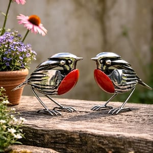 May include: Two metal robin sculptures with shiny silver bodies and bright red breasts stand on a wooden surface. The birds have detailed feather designs and are facing each other. The background includes potted plants and flowers.