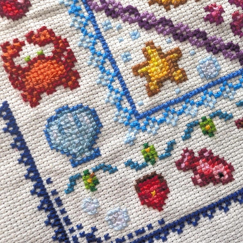 May include: Close-up of a cross-stitch embroidery featuring various marine life motifs. The design includes a crab, starfish, seashell, and other sea creatures in vibrant colors like orange, yellow, blue, and purple, stitched on white fabric.