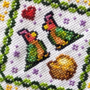 May include: Close-up of a cross-stitch design on white fabric. The design features two stylized birds, a red heart, and a shell, all within a green vine border. Additional small flowers and stars are scattered throughout the design.