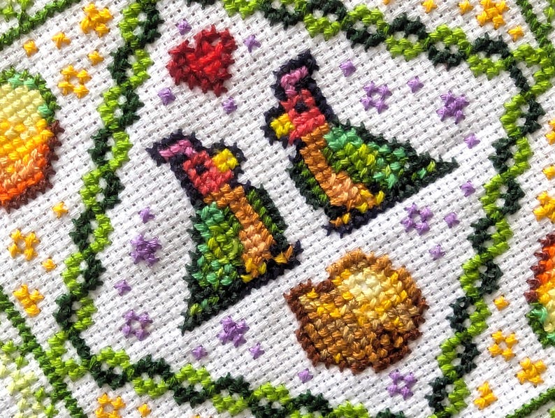 May include: Close-up of a cross-stitch design on white fabric. The design features two stylized birds, a red heart, and a shell, all within a green vine border. Additional small flowers and stars are scattered throughout the design.