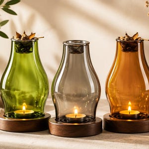 May include: Three glass candle holders with rounded bodies in green, gray, and amber. Each holder sits on a circular dark wood base and contains a lit tea light candle. The tops feature a metal insert holding dried botanical elements.