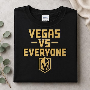 May include: A black short-sleeved t-shirt featuring the gold text "VEGAS -VS- EVERYONE" printed on the front. Below the text is a gold graphic of a knight's helmet shield logo. The shirt has a Gildan brand label at the inner neckline.