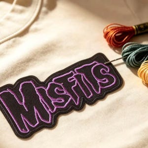 Może przedstawiać: Kremowy t-shirt z czarną, haftowaną naszywką z napisem MISFITS wyszytym fioletową nicią. W naszywkę wbita jest srebrna igła, a obok znajdują się trzy motki nici do haftowania w kolorach czerwonym, morskim i żółtym.