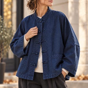 Puede incluir: Una chaqueta de mezclilla azul oscuro con cuello mao, mangas anchas de tres cuartos y un cierre frontal asegurado con cinco botones de palanca cubiertos de tela. La chaqueta incluye dos bolsillos de parche en la parte inferior frontal.