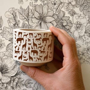 Pu&ograve; includere: Un piccolo vaso di ceramica bianco sporco con un motivo ripetuto di sagome di animali marroni, tra cui giraffe, mucche e conigli. Il vaso &egrave; cilindrico. Lo sfondo &egrave; un disegno floreale.