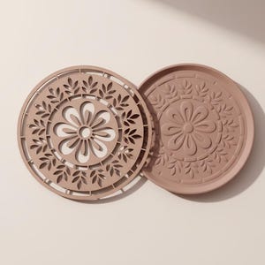 May include: Two circular coasters in a muted pink hue. One has a laser-cut floral design, featuring a central flower and leaves. The other has a raised floral pattern. Each coaster is approximately 10 cm in diameter.