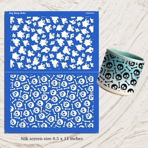 Puede incluir: Un esténcil de serigrafía azul con patrones de fantasmas y calaveras blancos, y una taza de cerámica con un diseño de calavera y huesos cruzados. El esténcil mide 21,6 x 27,9 cm. La taza tiene un interior azul claro.