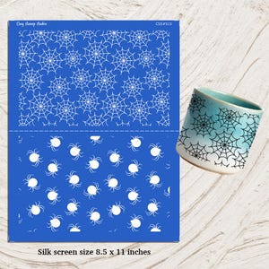 Puede incluir: Un esténcil de serigrafía azul con telarañas y arañas blancas, junto a una taza de cerámica con un diseño de telaraña. El esténcil está etiquetado como "Clay stamp studio" y "CSS#515". El tamaño del esténcil es de 21,6 x 27,9 cm.