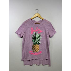 Könnte beinhalten: Ein kurzärmeliges T-Shirt in meliertem Lila mit einer Ananas-Grafik in der Mitte. Der Schriftzug Juicy Couture ist in leuchtendem Pink über und unter der Ananas aufgedruckt. Das Shirt hat einen asymmetrischen Saum mit Seitenschlitzen.