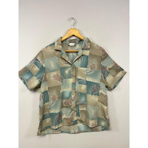 Op de afbeelding: Een overhemd met korte mouwen en knoopsluiting, voorzien van een patchworkpatroon van gedempte blauwgroene en beige vierkanten. Elk vierkant bevat een verfijnde bloemen-paisleyprint in roze en groene tinten. Het overhemd heeft een klassieke kraag.