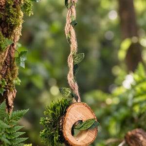 Può includere: Un ornamento sospeso composto da una fetta di legno circolare con bordo in corteccia naturale e un ciuffo di muschio verde. L'elemento in legno è appeso a una corda di juta intrecciata con piccole foglie verdi artificiali che risalgono.