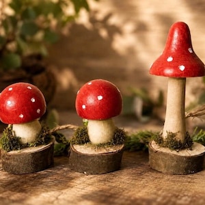 May include: Three decorative mushroom figurines with bright red caps featuring white spots. Each mushroom has a light-coloured stem and is mounted on a circular wooden base with green moss accents. The mushrooms vary in height and cap shape.