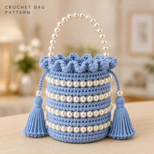 Può includere: Una borsa all'uncinetto azzurro chiaro con un manico di perle e quattro file orizzontali di perle bianche integrate nel corpo. La borsa presenta un bordo superiore arricciato e due coulisse laterali che terminano con nappe blu. Testo: CROCHET BAG PATTERN.