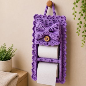 May include: A purple crocheted toilet paper holder with a scalloped edge and a decorative bow. The holder features a top flap secured by a wooden button and two open compartments, each containing a roll of white toilet paper. It hangs from a hook.