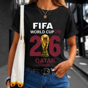 May include: Black t-shirt with the text "FIFA WORLD CUP 26" in white. The number "26" is in burgundy, with a gold trophy in the middle. Below the number is the word "QATAR" and the year "2026" in white.