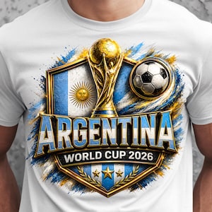 Puede incluir: Camiseta blanca con un diseño colorido de Argentina para la Copa del Mundo 2026. El diseño incluye la bandera argentina, un trofeo dorado, un balón de fútbol y la palabra "ARGENTINA" en azul. El diseño está enmarcado por un borde dorado.