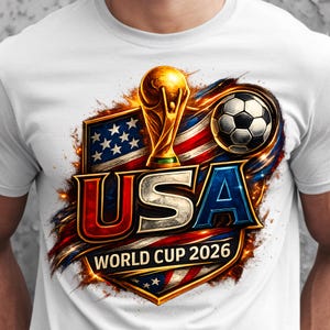 Puede incluir: Camiseta blanca con un gráfico colorido para la Copa del Mundo de 2026. El diseño incluye el emblema de EE. UU., un trofeo dorado, un balón de fútbol y el texto "USA WORLD CUP 2026" en rojo, blanco y azul.