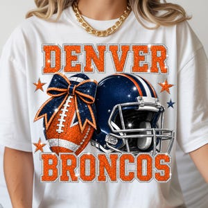 Puede incluir: Una camiseta blanca con un estampado gráfico que muestra el texto DENVER BRONCOS en purpurina naranja. El diseño incluye un balón de fútbol americano con un lazo azul marino brillante y un casco azul marino con rayas blancas.