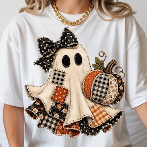 May include: A white short-sleeve t-shirt featuring a graphic of a ghost adorned with a black polka-dot bow. The ghost is decorated with various fabric patches in orange, black, and white patterns, including gingham and floral prints, next to a pumpkin.