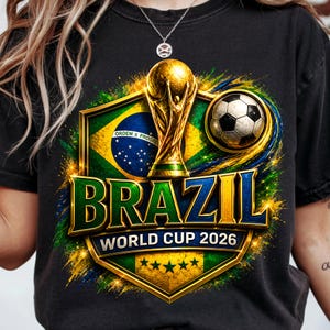 Puede incluir: Una camiseta negra con un gráfico de la bandera de Brasil, un trofeo dorado y un balón de fútbol. El texto dice BRAZIL WORLD CUP 2026. El diseño incluye un emblema de escudo con cinco estrellas verdes y salpicaduras amarillas y verdes.