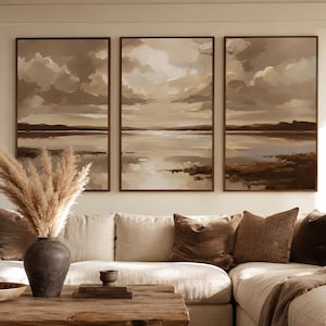 May include: A living room scene featuring a sectional sofa with neutral-toned cushions and decorative throw pillows in shades of brown. Above the sofa, three framed landscape paintings depict a serene waterscape with a cloudy sky. A vase of pampas grass sits on a wooden coffee table.