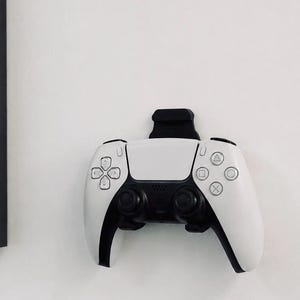 May include: A white and black wireless video game controller mounted on a wall using a black plastic bracket. The controller features a central touchpad, two analogue sticks, a directional pad, and four action buttons with triangle, circle, cross, and square symbols.