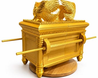 Op de afbeelding: Een goudkleurige rechthoekige kist met twee gevleugelde figuren op het deksel die naar elkaar toe zijn gericht. De kist heeft decoratieve zuilen op de hoeken, een gestructureerde rand en twee lange horizontale stangen aan de zijkanten.