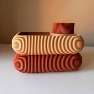 May include: A two-tiered storage tray with a ribbed texture. The top section is light peach with a dark terracotta cylindrical cup attached, while the bottom section is a solid dark terracotta oval base with rounded edges.