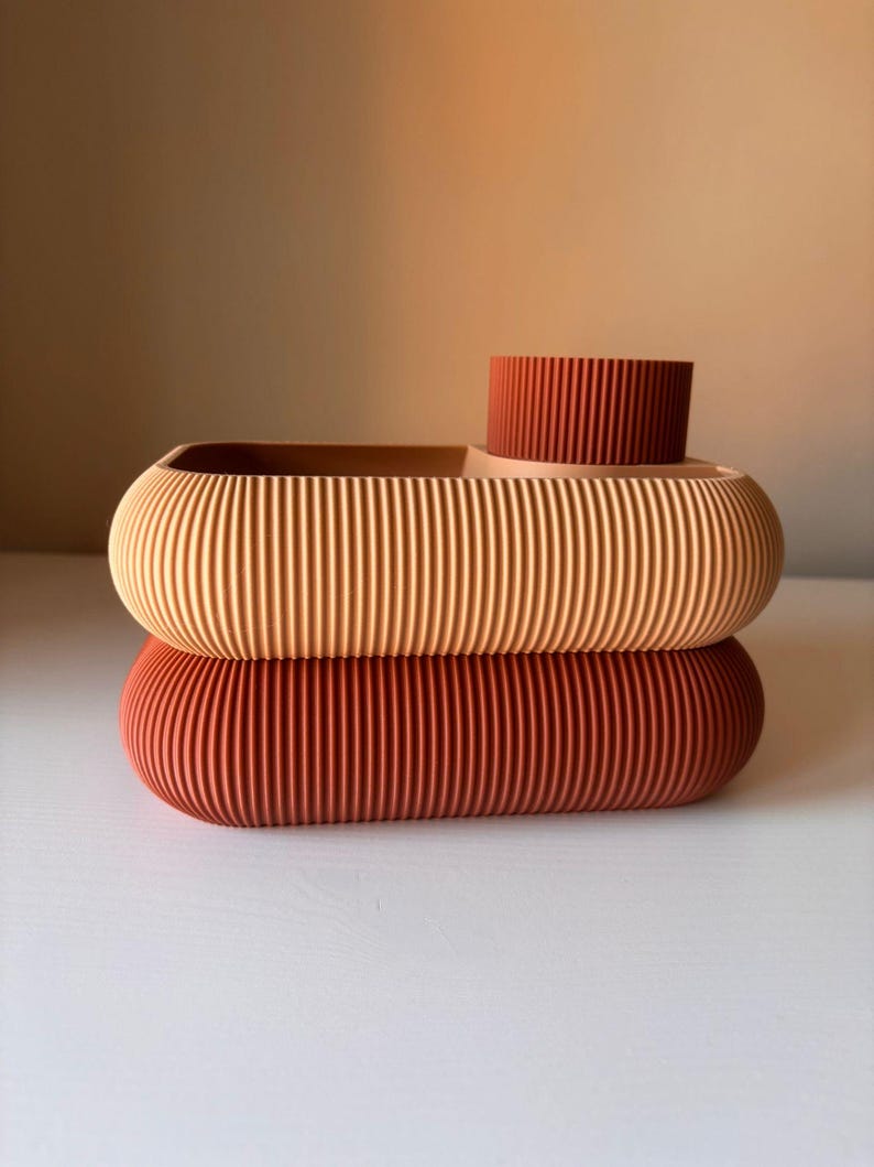 May include: A two-tiered storage tray with a ribbed texture. The top section is light peach with a dark terracotta cylindrical cup attached, while the bottom section is a solid dark terracotta oval base with rounded edges.