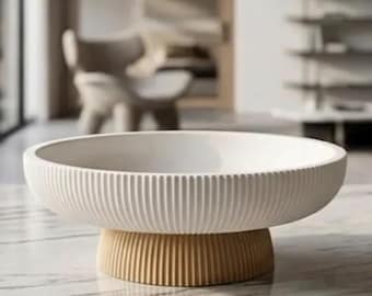 Op de afbeelding: Een ondiepe, ronde witte kom met een verticaal geribbelde buitenkant. De kom rust op een contrasterende, kortere, verticaal geribbelde voet in een warme beige of lichtbruine kleur, wat zorgt voor een tweekleurig uiterlijk.