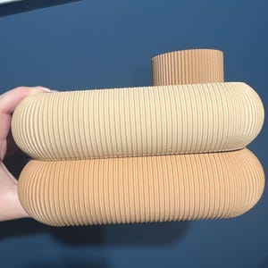 May include: A beige ceramic vase featuring a stacked, rounded design with a ribbed, textured surface. The object consists of two horizontal, pill-shaped sections and a smaller, cylindrical opening at the top, all in a uniform light tan color.