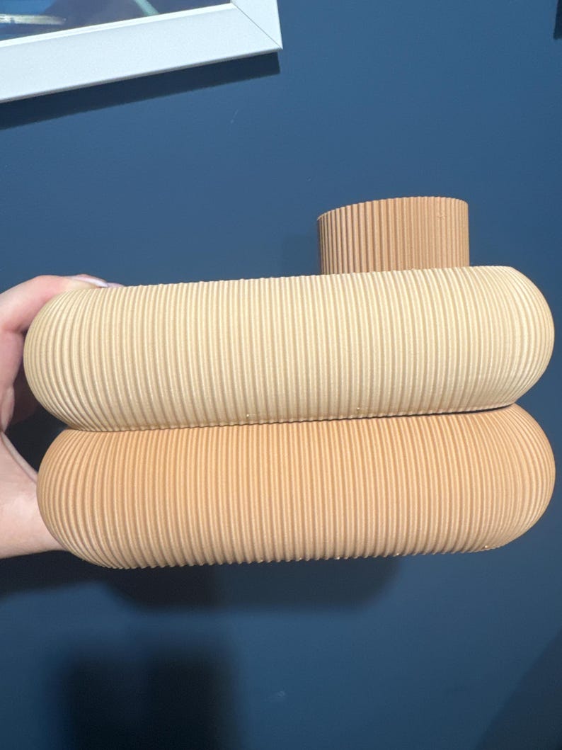 May include: A beige ceramic vase featuring a stacked, rounded design with a ribbed, textured surface. The object consists of two horizontal, pill-shaped sections and a smaller, cylindrical opening at the top, all in a uniform light tan color.
