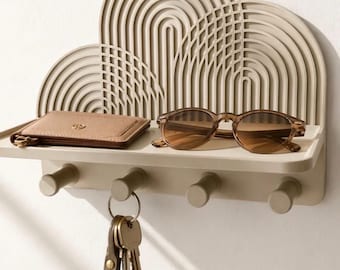 Op de afbeelding: Een beige wandplank met een decoratief achterpaneel met concentrische boogpatronen. De plank heeft vier cilindrische haken aan de onderkant waaraan een sleutelbos hangt, terwijl op de plank zelf een portemonnee en een zonnebril liggen.