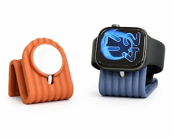 Op de afbeelding: Een geribbelde oplaadstandaard van terracotta-kleurige siliconen met een witte ronde magnetische oplader en een geribbelde oplaadstandaard van leisteenblauwe siliconen met een zwarte smartwatch die de tijd 17:42 digitaal weergeeft.