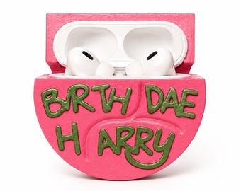 Op de afbeelding: Een felroze, halfronde beschermhoes voor draadloze oordopjes, met de handgeschilderde groene tekst "BRTH DAE HARRY" op de voorzijde. De hoes staat open en toont een witte binnenkant met een paar witte draadloze oordopjes erin.