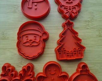 Op de afbeelding: Een set van zeven rode plastic koekjesvormpjes met kerstthema, waaronder een sneeuwpop, een persoon met een bril, de Kerstman, een kerstboom met cadeautjes, een rendier, een peperkoekmannetje en een ronde kerstbal.