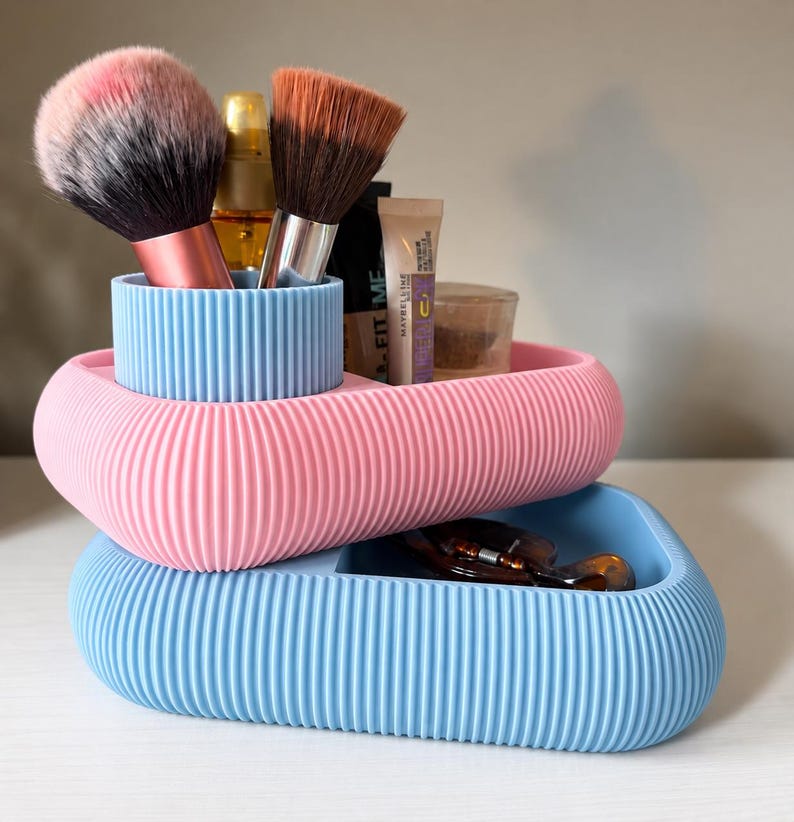 May include: A stacked two-tier desk organizer with a ribbed texture. The top tier is pink with a light blue cylindrical brush holder, and the bottom tier is light blue. Various makeup brushes and cosmetic tubes are stored inside the compartments.
