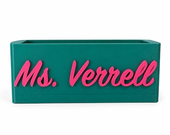 Op de afbeelding: Een rechthoekige, blauwgroene bureau-organizer met de naam Ms. Verrell in verhoogde, felroze cursieve letters op de voorzijde. De houder heeft een effen, matte afwerking en een open bovenkant, ontworpen voor het opbergen van kantoorbenodigdheden.