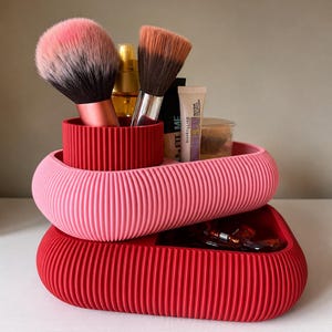 May include: A two-tiered, ribbed storage organizer featuring a pink oval top tray and a red oval base tray. The top tray holds two makeup brushes with pink and brown bristles, a gold bottle, and a Maybelline Fit Me tube.