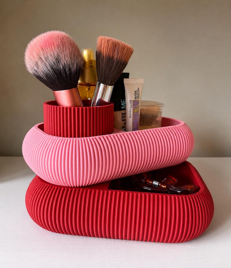 May include: A two-tiered, ribbed storage organizer featuring a pink oval top tray and a red oval base tray. The top tray holds two makeup brushes with pink and brown bristles, a gold bottle, and a Maybelline Fit Me tube.