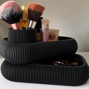 May include: A black, ribbed, two-tiered cosmetic organizer with a rounded rectangular shape. The top tier features a cylindrical brush holder containing two makeup brushes, while the lower tier holds various makeup tubes and a small container.