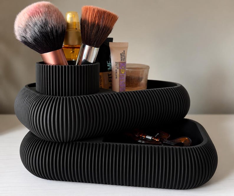 May include: A black, ribbed, two-tiered cosmetic organizer with a rounded rectangular shape. The top tier features a cylindrical brush holder containing two makeup brushes, while the lower tier holds various makeup tubes and a small container.