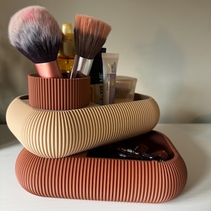 May include: A two-tiered, ribbed storage organizer in terracotta and beige. The top tier holds two makeup brushes with pink and brown bristles, a gold bottle, and a Maybelline Fit Me tube. The bottom tier contains a dark hair accessory.