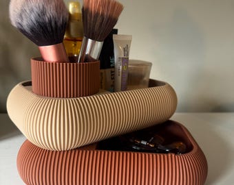 Op de afbeelding: Een geribbelde opbergorganizer met twee niveaus in terracotta en beige. Het bovenste niveau bevat twee make-upkwasten met roze en bruine haren, een gouden flesje en een Maybelline Fit Me tube. Het onderste niveau bevat een donker haaraccessoire.