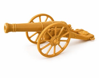 May include: A tan-colored plastic toy cannon featuring a long barrel, two large spoked wheels, and a trailing carriage frame. The design includes a circular emblem on the rear section of the carriage, with a hollow muzzle and detailed axle hubs.