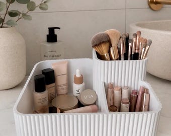 Op de afbeelding: Een witte, geribbelde make-up organizer van kunststof met meerdere vakken. De organizer bevat diverse cosmetica, waaronder make-up kwasten, foundationflesjes, lippenstiften en huidverzorgingsproducten met zichtbare labels zoals Ilia en Laura Mercier.